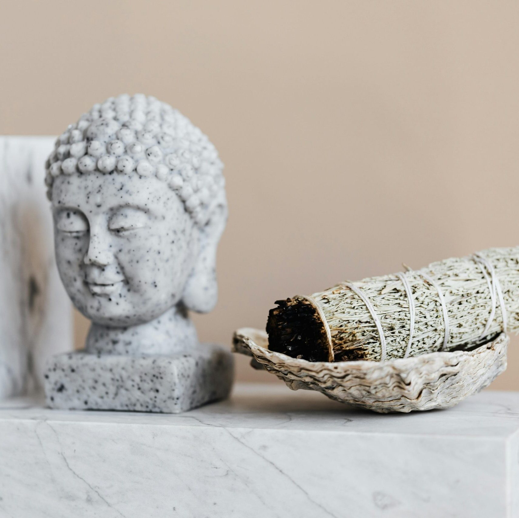 Bust of Tibetan Buddha with closed eyes made of grey speckled marble near decorative plate with sage incense bundle on beige background
