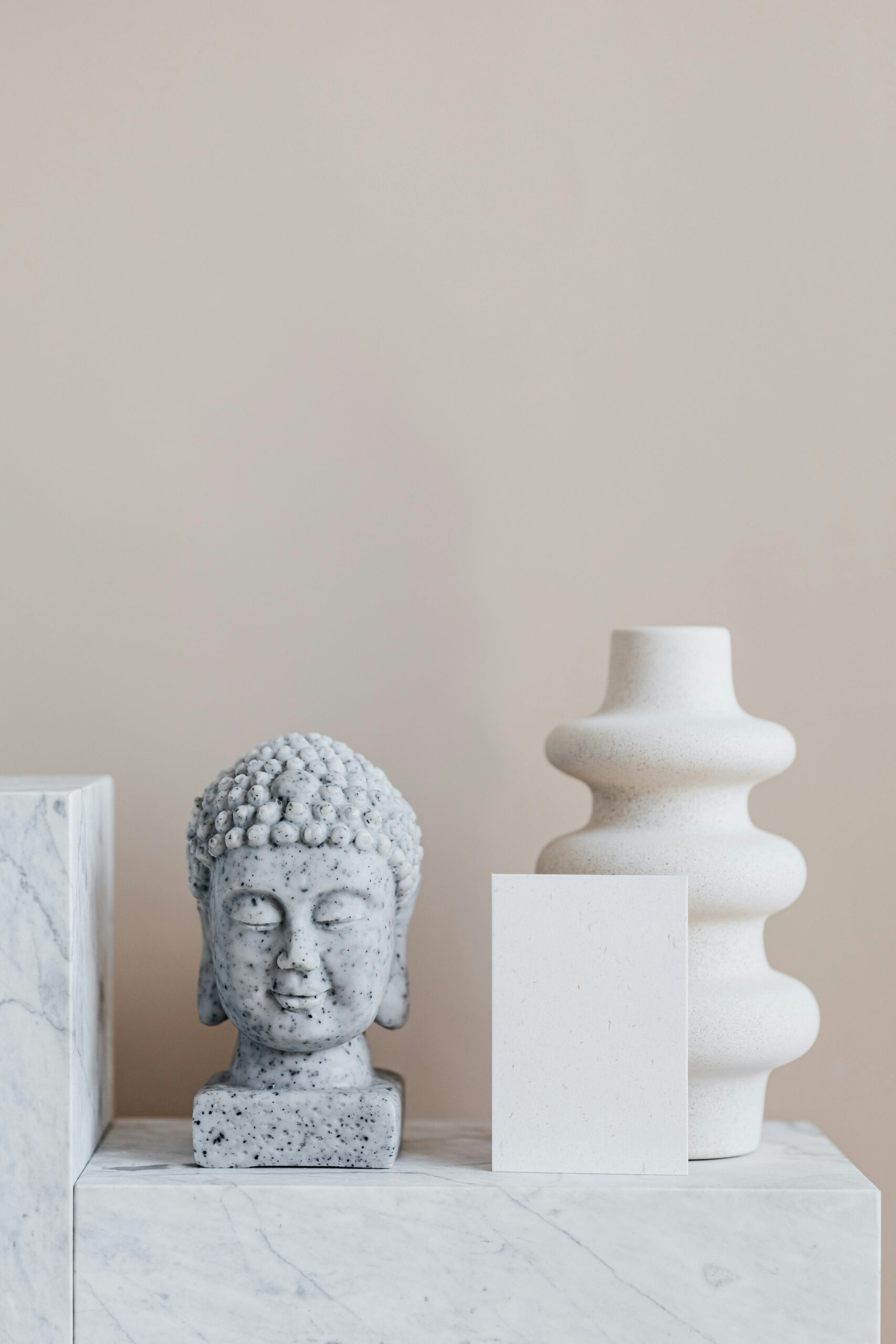 Granite bust of Buddha placed near white ceramic vase of creative geometric shape and empty card on white marble stand against beige wall